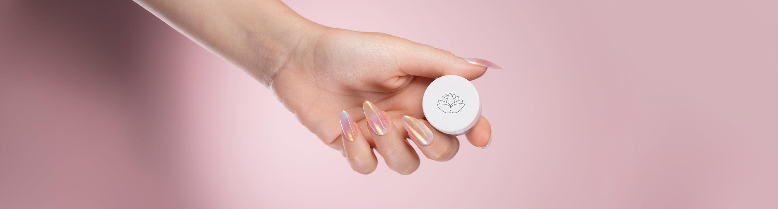 Mano con uñas de gel iridiscentes sosteniendo envase blanco de manicura sobre fondo rosa.