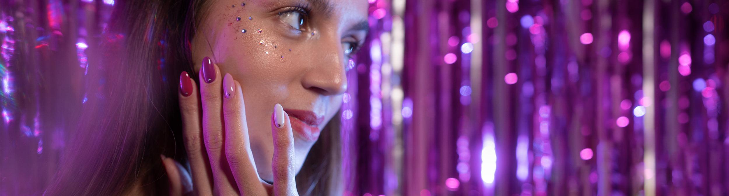 Uñas decoradas en tonos rosa y lila, maquillaje con brillos, fondo festivo violeta
