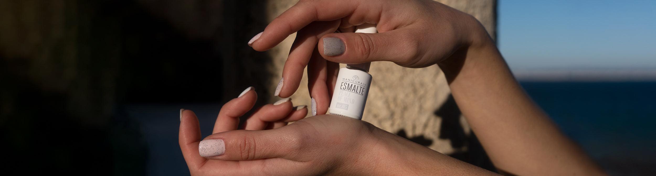 Manos con manicura y esmalte de uñas blanco en botella, fondo exterior con mar y piedra