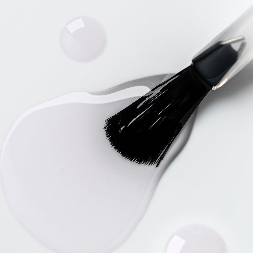 Brocha de manicura aplicando gel transparente sobre superficie blanca, detalle de pincel y brillo